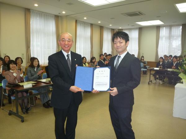 受賞者が座って拍手をしている中で、競技かるた日本一の岸田名人と市長がふるさと大使委嘱状を手に持った写真